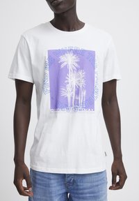 Camiseta de algodón blanca con un gráfico morado, palmeras blancas y texto. Manga corta y cuello redondo. Corte casual.