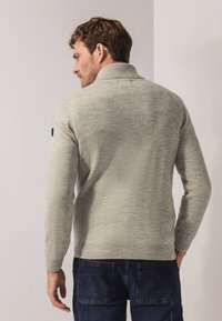 Hellgrauer Strickpullover mit hohem Kragen, gerippten Ärmelbündchen und Saum, der eine subtile Textur aufweist. Kombiniert mit dunkelblauen Jeans.