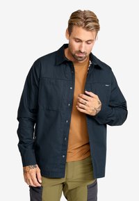 Mörkblå button-up skjorta med två bröstfickor, lång ärm och en strukturerad krage, lagd över en brun skjorta. Texturerat tyg.