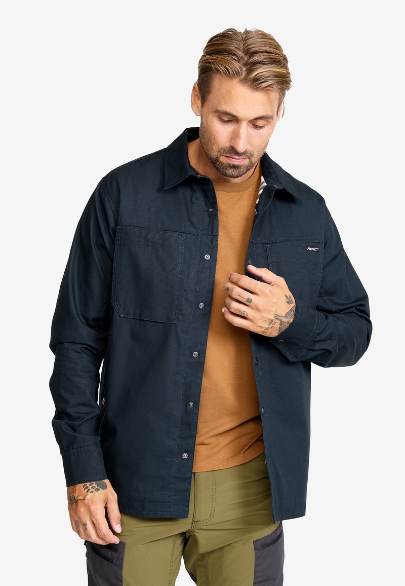 Mörkblå button-up skjorta med två bröstfickor, lång ärm och en strukturerad krage, lagd över en brun skjorta. Texturerat tyg.