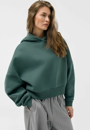 Kobieta o długich włosach, ubrana w ciemnozieloną, oversize'ową bluzę z kapturem oraz szare szerokie spodnie w prążki, z rękami w kieszeniach, patrząca w prawo.