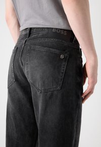 Los jeans de mezclilla negros presentan un acabado desvanecido, dos bolsillos traseros con costuras distintivas y una etiqueta de logo en la cinturilla.