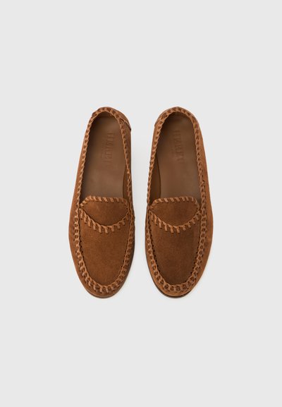 Καφέ μοκασίνια από σουέντ με ραμμένο σχέδιο κατά μήκος των άκρων και λείο δερμάτινο πάτο. Το επάνω μέρος διαθέτει στρογγυλεμένη μύτη σε στυλ μοκασινιού.