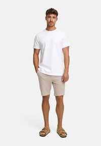 Hombre de pie con las manos en los bolsillos de unos pantalones cortos beige, vistiendo una camiseta blanca simple y sandalias marrones sobre un fondo blanco.
