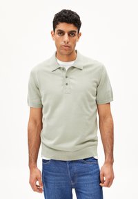 Lichtgroene polo met een geribbelde kraag en korte mouwen, voorzien van een drieknoopsluiting en een aansluitende zoom. Gdragen over een wit shirt.