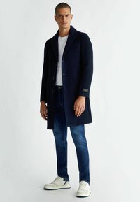 Marineblauwe wollen blended jas met omslagrevers, knoopsluiting en twee voorzakken, gecombineerd met blauwe denim jeans en witte sneakers.