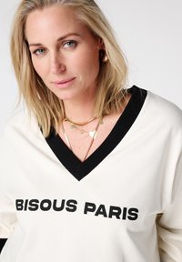 Roomkleurige pullover met zwarte bies, voorzien van een V-hals ontwerp en gedurfde tekst "BISOU PARIS". Draagt meerdere gouden kettingen in laagjes.