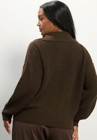 Brunstickad tröja med ribbad textur, med en bred krage och avslappnad passform, visad bakifrån. Mjuk tyg med långa ärmar.