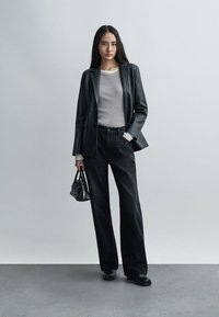 Γυναίκα όρθια φορώντας μαύρο δερμάτινο σακάκι, γκρι ριμπ πλεκτή μπλούζα, μαύρο παντελόνι με φαρδιά πόδια, μαύρες μπότες, κρατώντας μικρή μαύρη τσάντα χειρός.