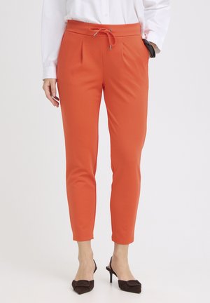Mujer que lleva pantalones tobilleros ajustados de color naranja con cintura de cordón, combinados con camisa blanca y zapatos de tacón negros de punta afilada.