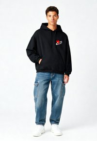 Felpa nera con cappuccio e tasca frontale, con un grafiche a forma di cuore rosso e spilla di sicurezza. Indossata con jeans cargo azzurri chiari e sneakers bianche.