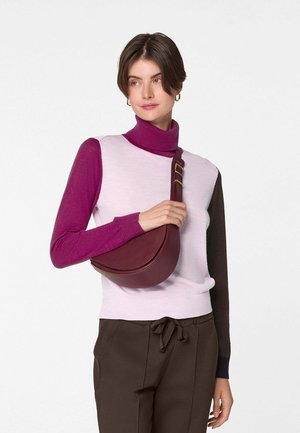 Femme portant un pull à col roulé bicolore et un pantalon marron, tenant un sac bandoulière demi-lune bordeaux sur son épaule.