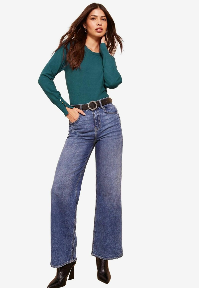 Maglione verde a maniche lunghe con dettagli di bottoni, abbinato a jeans a vita alta blu a gamba larga e stivali neri con tacco. Sfondo neutro.
