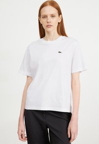 Weißes Baumwoll-T-Shirt mit Rundhalsausschnitt, kurzen Ärmeln und grünem Lacoste-Logo, das auf der linken Brust aufgestickt ist. Gerade Passform.