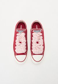Červené plátěné tenisky s bílými gumovými podrážkami, s růžovými kostkovanými tkaničkami a klasickým brandingem Converse All Star na jazyku.