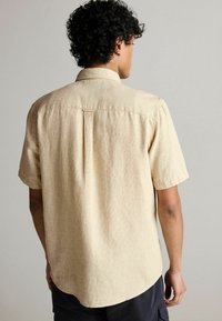 Camisa de manga corta color beige con una tela texturada, cuello abotonado y un pliegue en la parte trasera, que presenta un dobladillo curvado y un corte relajado.
