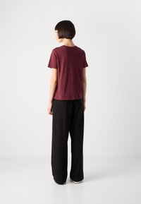 Woman with short dark hair standing facing away, wearing a burgundy short-sleeve shirt and black wide-leg pants against a plain background.