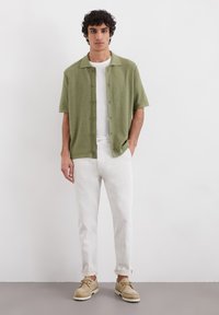 Olive green short-sleeve shirt, white t-shirt underneath, light-colored rolled-up trousers, and beige shoes on a neutral background.