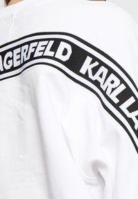 Bílý svetr s černým pruhem přes záda, na kterém je tučně napsáno "KARL LAGERFELD" v bílé barvě. Materiál vypadá jemně a texturovaně.