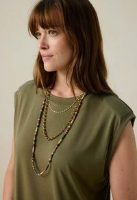 Haut vert olive à manches courtes, superposé avec trois colliers : une chaîne en or et des fils de perles colorées, mettant en valeur le design du décolleté.