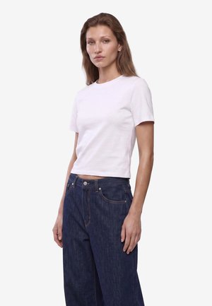 Giovane donna con lunghi capelli castani che indossa una maglietta bianca semplice a maniche corte e jeans a vita alta blu scuro su sfondo bianco.