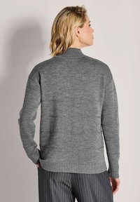 Frau mit kurzem blonden Haar, die einen grauen Strickpullover und graue Nadelstreifen-Hose trägt, steht mit dem Rücken zur Kamera.