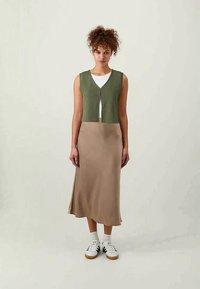 Πράσινο πλεκτό γιλέκο με κουμπί μπροστά, φορεμένο πάνω από ένα κρεμ μπλουζάκι και μία μπεζ φούστα slip, συνδυασμένο με λευκά αθλητικά παπούτσια. Απαλή υφή υφάσματος.