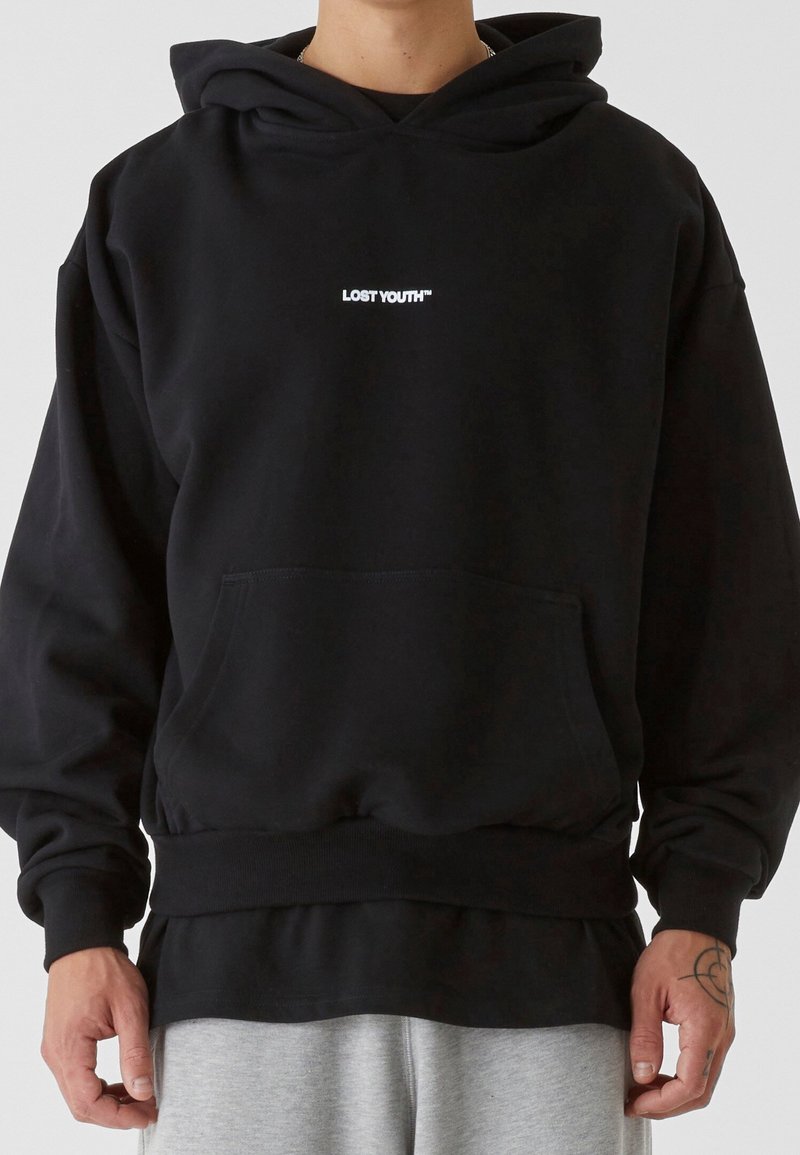 Sweat à capuche noir avec le texte "LOST YOUTH™" sur la poitrine, porté par une personne avec un tatouage visible sur la main droite, assorti à un pantalon gris clair.