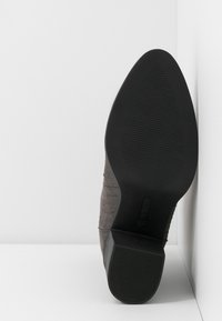 Botte à bout pointu à semelle noire avec talon bloc, semelle texturée antidérapante, positionnée debout contre des murs blancs d'angle.