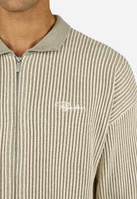 Lengvas zip-up megztinis su nerti tekstūra, turintis pakaitomis šviesiai rudos ir pilkos spalvų juostas. Įeina pilkas apykaklė ir išsiuvinėtas emblemą.