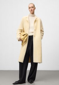 Vrouw in een crèmekleurige wollen jas, licht coltruitje, zwarte wijde broek, zwarte puntige ballerina's en houdt een beige handtas vast.