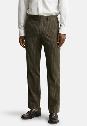 Homme portant un pantalon à fines rayures olive foncé, une chemise blanche habillée et des mocassins en cuir marron foncé, debout devant un fond uni.