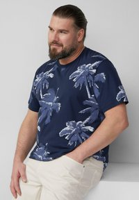 Granatowa koszulka z białym wzorem palm, okrągły dekolt, krótki rękaw, wykonana z miękkiego materiału, zestawiona z jasnymi spodniami.