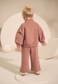 Completo due pezzi color rosa polvere con un pullover dalle maniche voluminose e pantaloni dalla vestibilità rilassata. Il materiale sembra morbido con una texture liscia.