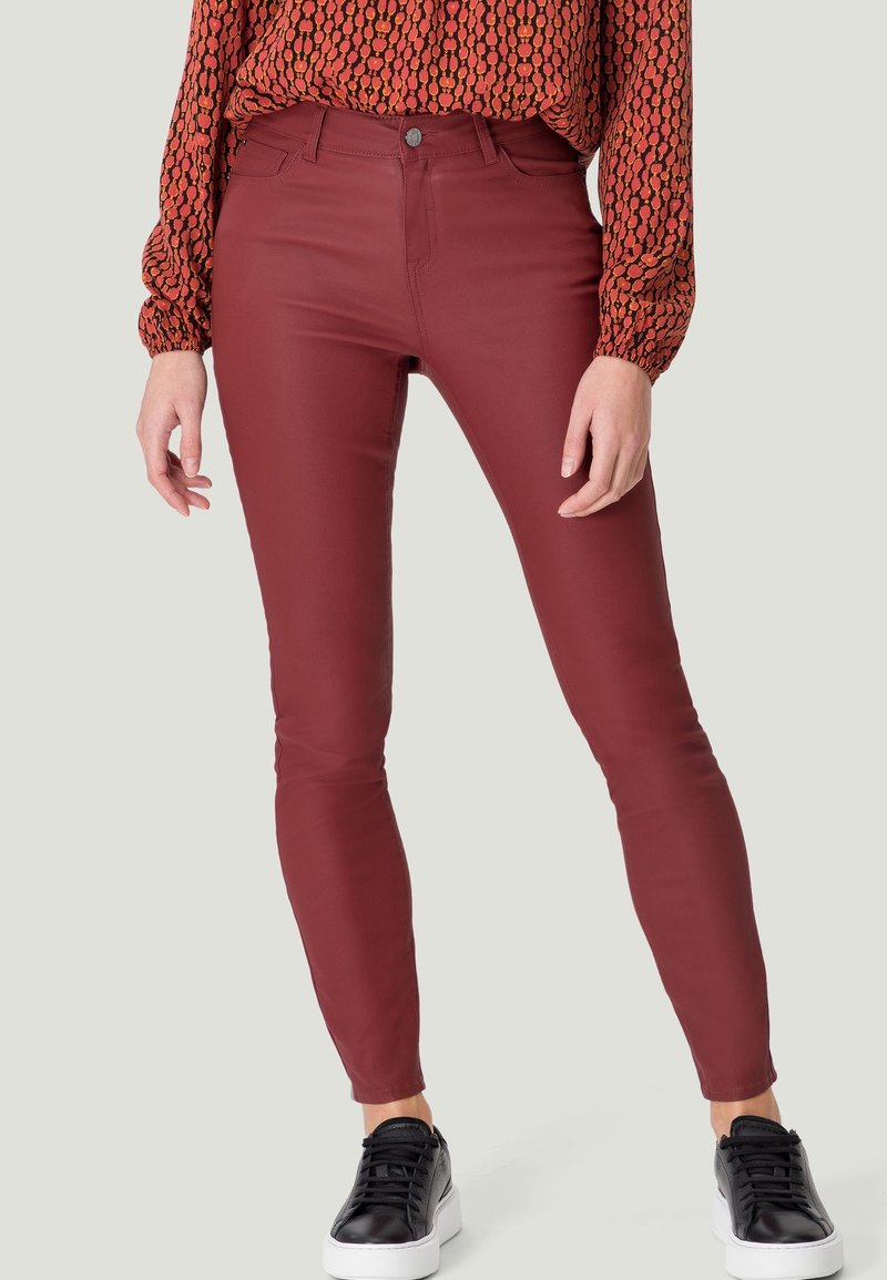 Femme portant un pantalon rouge foncé ajusté, un haut à manches longues à motifs rentré, et des baskets noires aux semelles blanches.