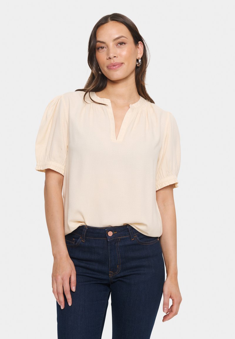 Blusa a maniche corte di color crema in tessuto leggero, con scollatura a V e maniche a sbuffo arricciate, abbinata a jeans di colore blu scuro.
