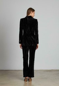 Completo in velluto nero testurizzato con blazer su misura con colletto alto e pantaloni aderenti, abbinato a tacchi a punta.