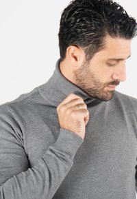 Pull à col roulé gris en texture côtelée douce, avec des manches longues et une coupe ajustée, porté avec une main tenant le col.