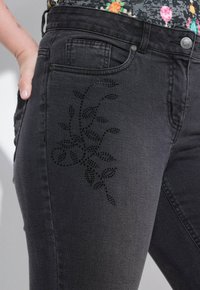 Los pantalones de mezclilla negros presentan un diseño floral hecho de pequeños strass en el muslo. Incluyen bolsillos estándar y un cierre de botón.