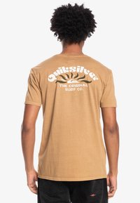 Hnědé tričko s krátkým rukávem a volným střihem. Na zadní straně se nachází grafické logo v bílé barvě s motivem slunce a vln. Jednoduchá, měkká textura.