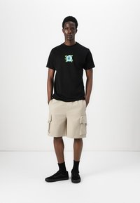 Sort kortærmet t-shirt med blå blomstermønster på brystet, parret med beige cargo shorts med sidelommer. Sorte sneakers og sokker.