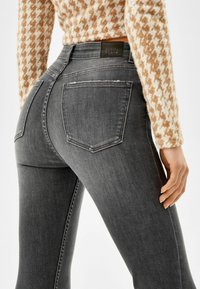 Pantalones vaqueros ajustados de mezclilla gris con cintura alta, que presentan bolsillos traseros y un ligero desgaste en los bordes. Etiqueta del logo en la cinturilla.