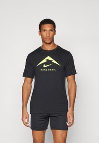 Svart kortermet T-skjorte med gult Nike Trail-logo og trekantet design. Laget av mykt stoff, kombinert med svarte shorts.
