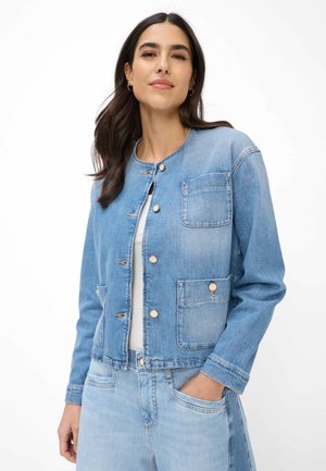 Vrouw met donker haar die een lichtblauwe denimjas met knopen draagt over een wit shirt, met één hand in de zak van bijpassende denim broek.
