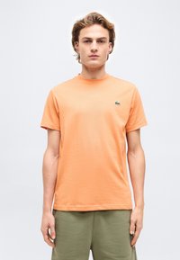 Νεαρός άνδρας που φοράει απλό πορτοκαλί κοντομάνικο T-shirt με μικρό πράσινο λογότυπο κροκοδείλου και χακί παντελόνι, στέκεται μπροστά σε φωτεινό φόντο.