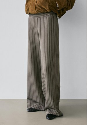 Osoba nosící široké kalhoty s žebrovanou strukturou v barvě taupe a vysokým pasem, v kombinaci s černými botami s špičatou špičkou a hnědým sakem.