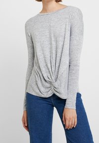 Haut gris à manches longues avec un détail de nœud torsadé à la taille, en tissu texturé. Associé à un jean denim bleu.