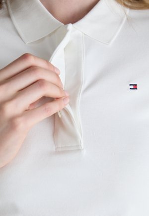 Hvit polo-skjorte med teksturert stoff. Har krage og en liten brodert logo nær brystet. Detalj med glidelås er synlig.