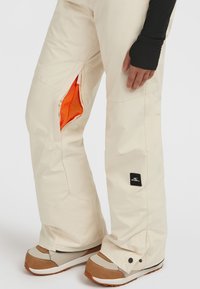 Beige sneeuwbroeken met mesh-voering aan de zijkanten, een zwart logo-patch en verstevigde enkels, gecombineerd met bruine en beige sneeuwlaarzen.