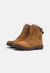 Bottines en suede marron avec des semelles en caoutchouc, des œillets en métal et une doublure en fourrure. Présente un logo circulaire sur le côté et un col rembourré.